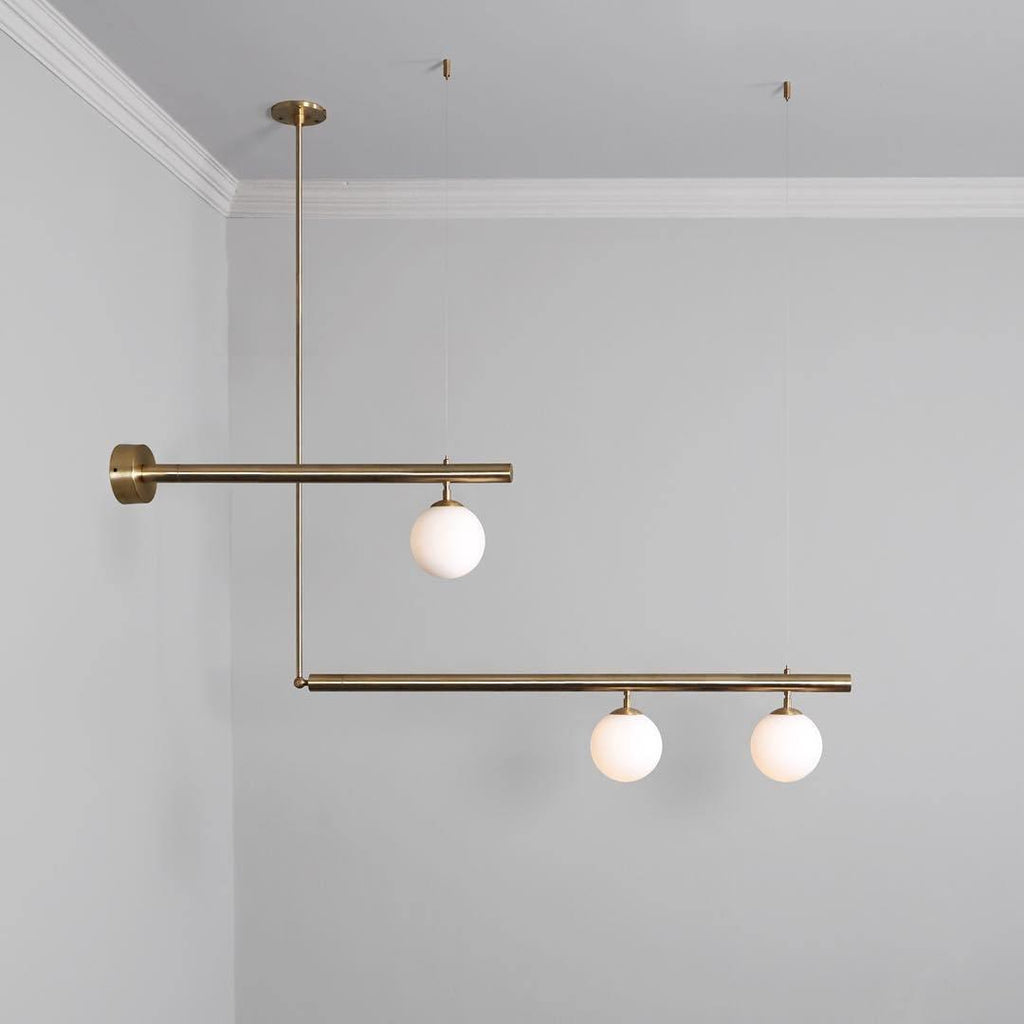 Clérachic gold pendant with offset bars and three globes, one arm fixed to a wall plate, against a light grey wall.