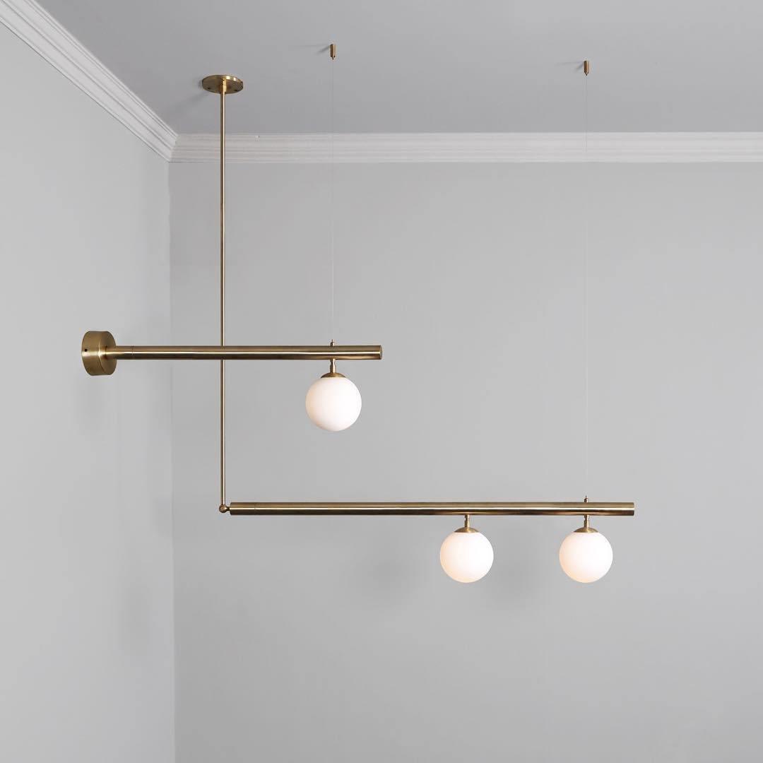 Clérachic gold pendant with offset bars and three globes, one arm fixed to a wall plate, against a light grey wall.