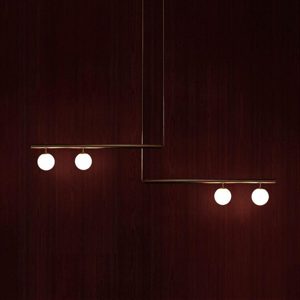Clérachic gold pendant with two offset bars and four lit globes, hanging against a deep red mahogany wall.