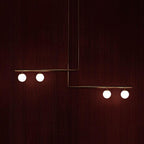 Clérachic gold pendant with two offset bars and four lit globes, hanging against a deep red mahogany wall.