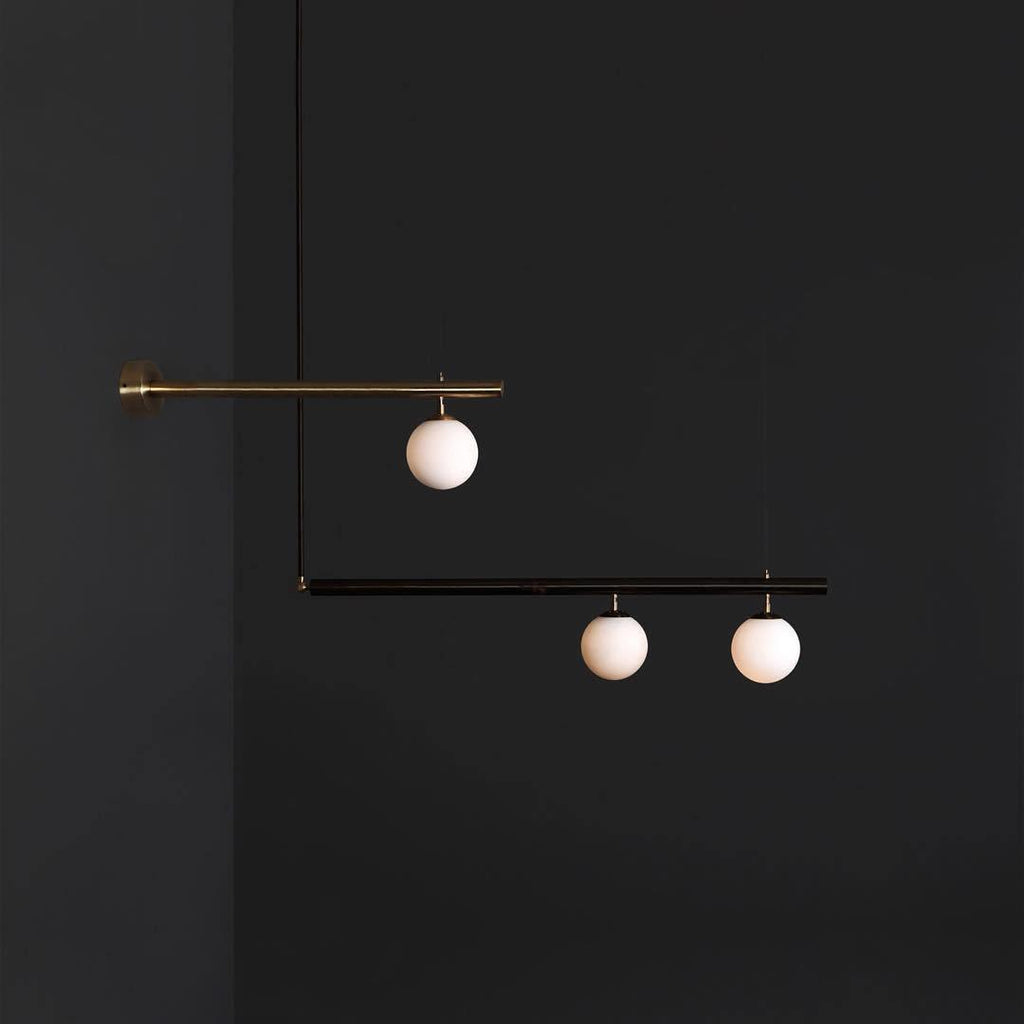 Clérachic pendant with a wall-fixed brass arm and a black bar, three lit globes in all, against a dark grey wall.