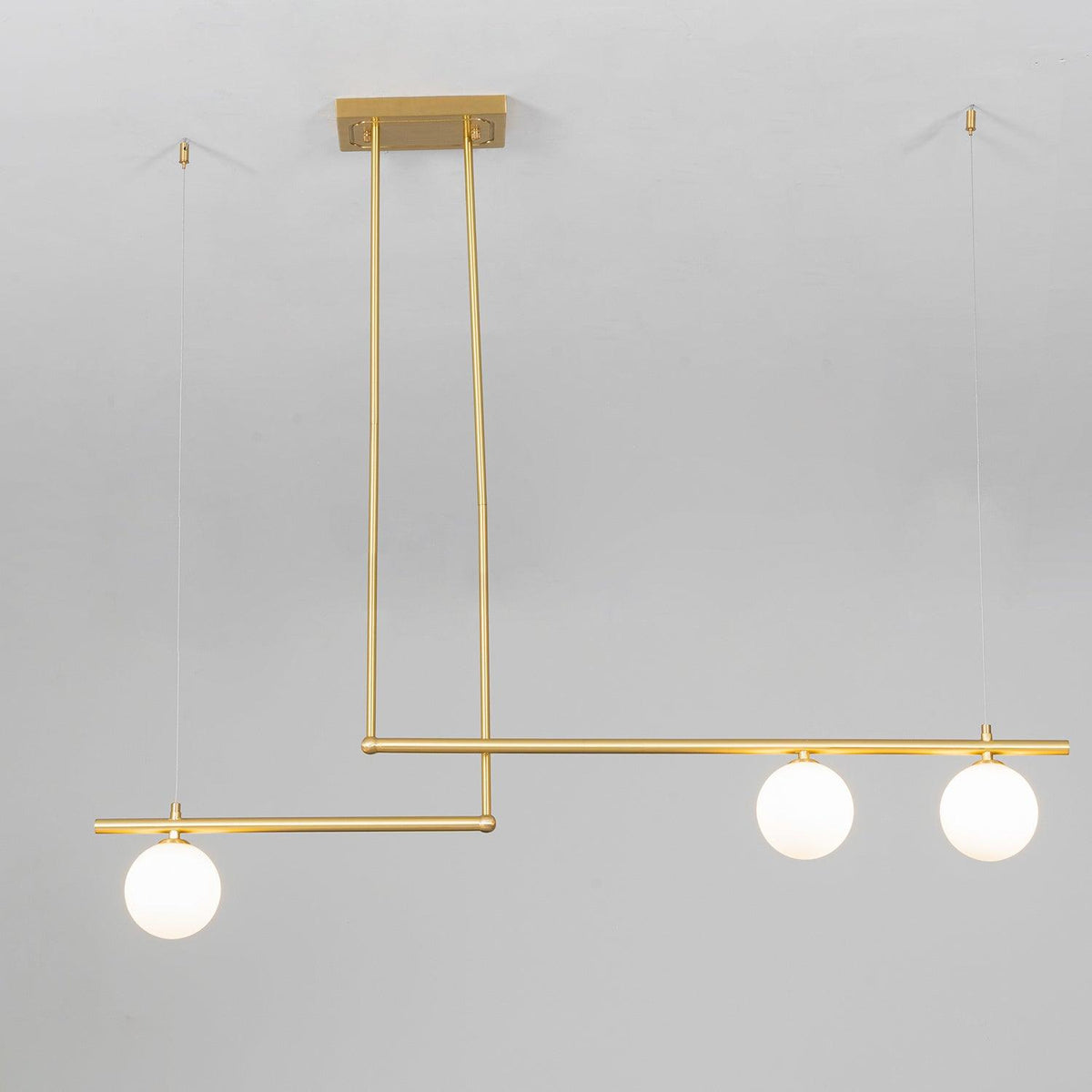 Close view of a three-globe Clérachic gold pendant hanging from its rectangular ceiling plate against a pale wall.