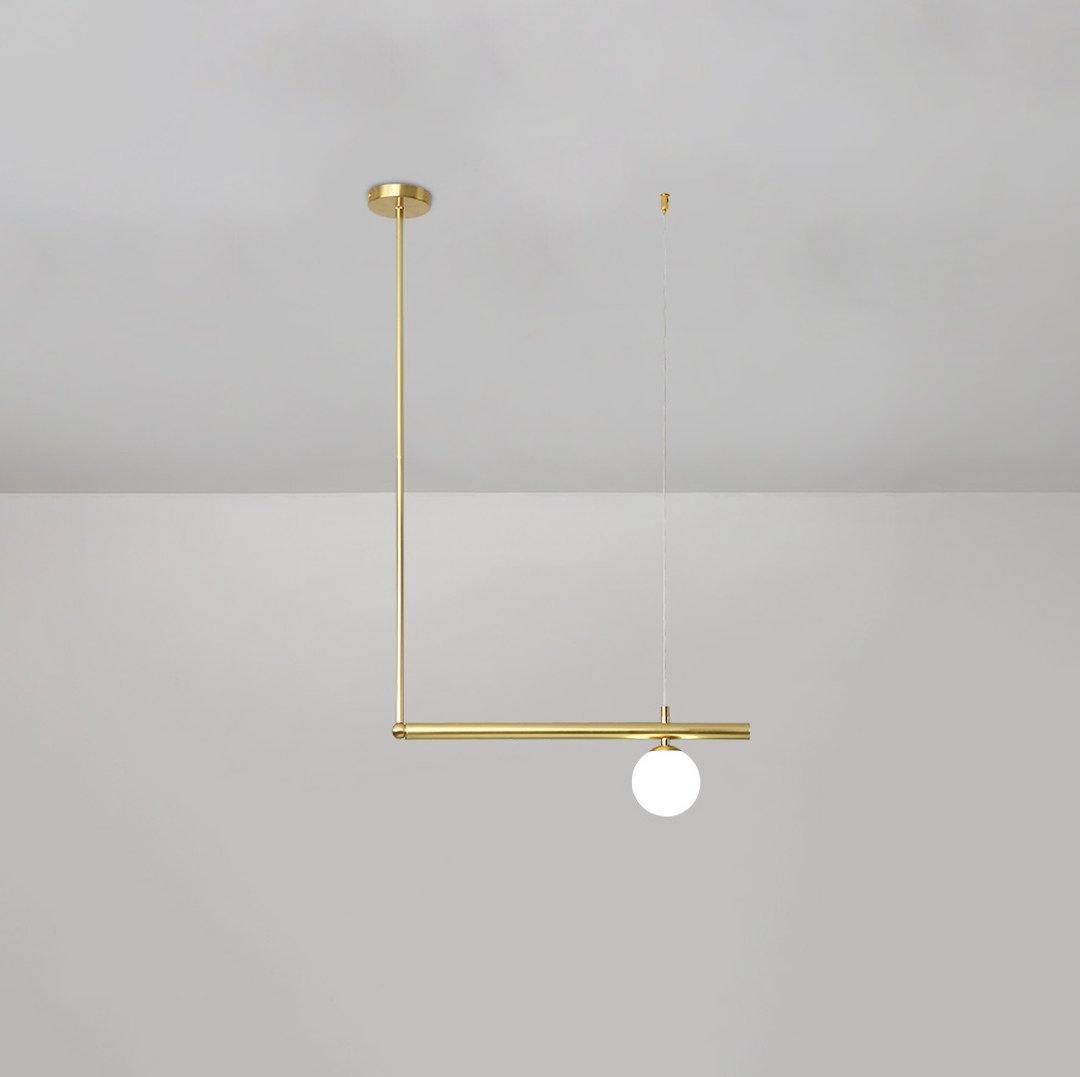Single-globe Clérachic pendant hung from a round gold ceiling cap, with a gold rod and a short bar, on a pale grey wall.