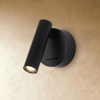 Clérachic matte black wall reading light, its cylinder head angled down and glowing warm beside a round backplate.