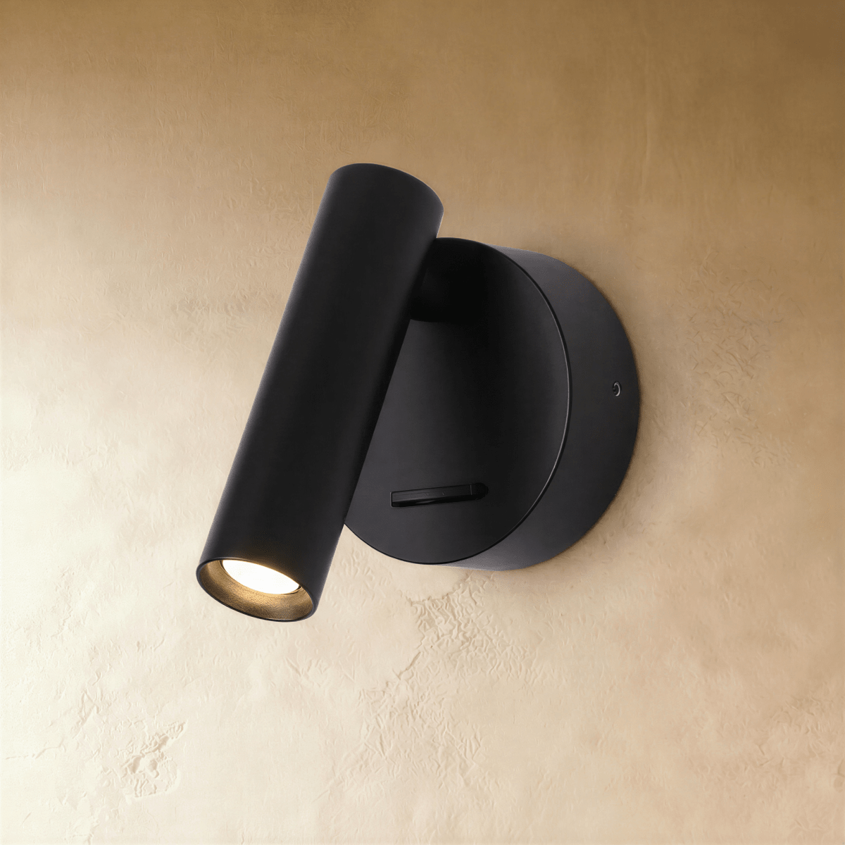 Clérachic matte black wall reading light, its cylinder head angled down and glowing warm beside a round backplate.