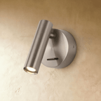 Clérachic brushed nickel wall reading light with a satin cylinder head and round backplate, lit warm on a plaster wall.