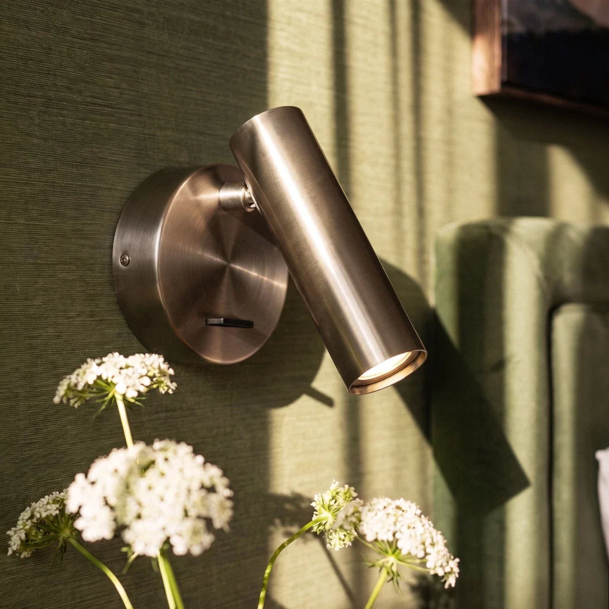 Clérachic satin metal wall reading light close up, its cylinder head aimed down over white wildflowers on a green wall.
