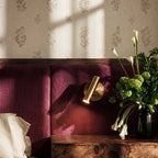 Clérachic brass wall reading light close up on a plum headboard, its cylinder head glowing beside a vase of white lilies.