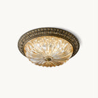 This Clérachic flush-mount ceiling light on a plain white backdrop, a lit amber pleated glass bowl in a leaf-patterned antique brass rim.