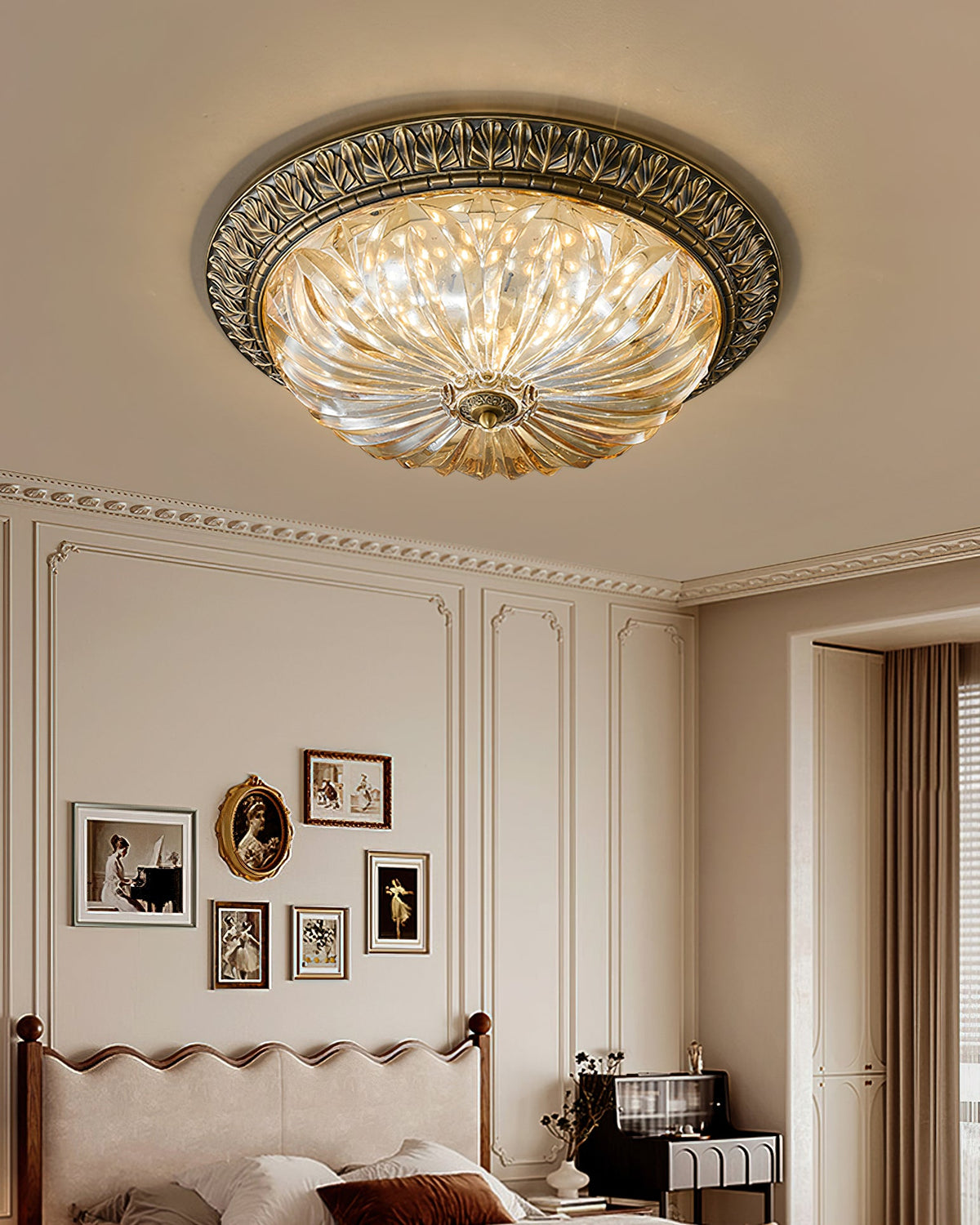 A Clérachic ceiling light in a tall panelled room, the amber glass lit above a scalloped headboard and a gallery of small frames.