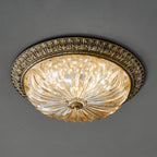 Lit Clérachic ceiling light seen at an angle on a grey ceiling, the ribbed amber glass glowing inside its antique brass rim.