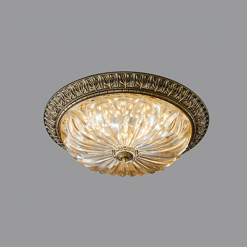 This Clérachic flush-mount ceiling light, lit, on a plain grey backdrop, its amber pleated glass in an antique brass rim.
