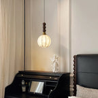 A small lit Clérachic pendant with a rounded, ribbed cream shade hangs by a curtain, beside a black writing desk and an angel figure.