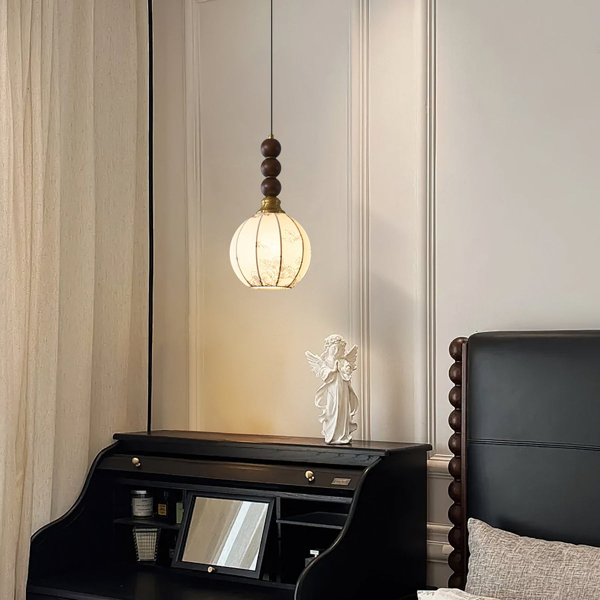 A small lit Clérachic pendant with a rounded, ribbed cream shade hangs by a curtain, beside a black writing desk and an angel figure.