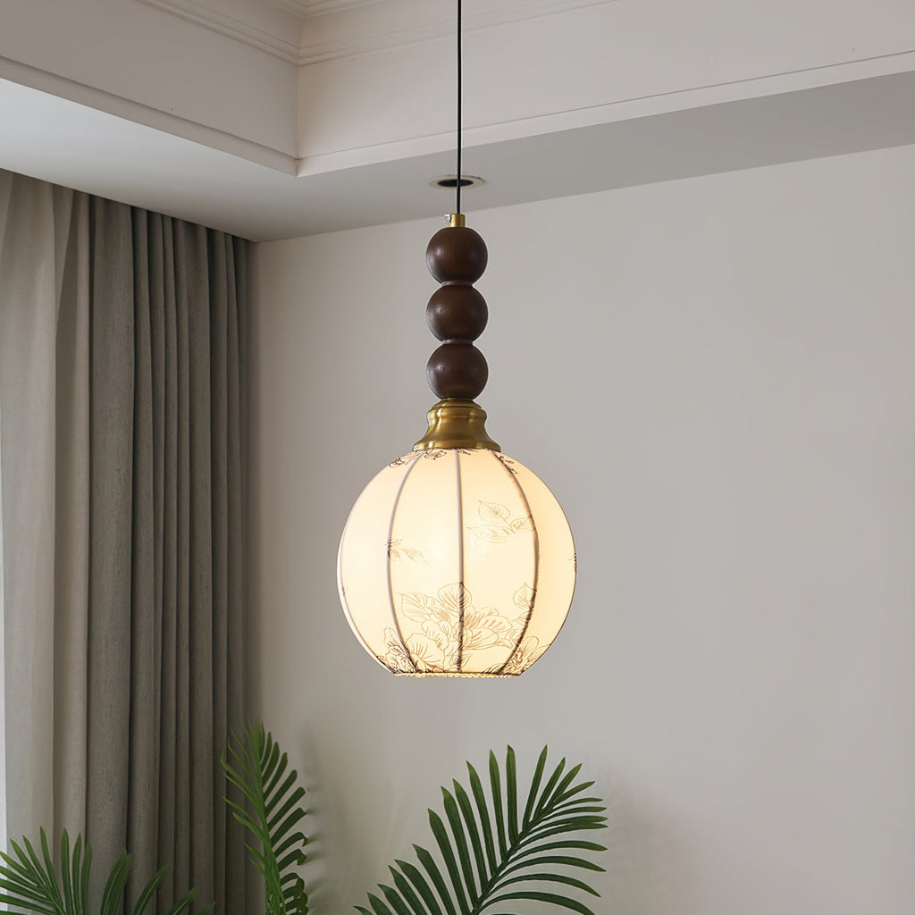 This Clérachic pendant has a round cream shade with fine ribs and a column of three walnut beads, lit warm beside a curtain and a palm.