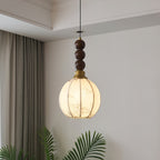 This Clérachic pendant has a round cream shade with fine ribs and a column of three walnut beads, lit warm beside a curtain and a palm.