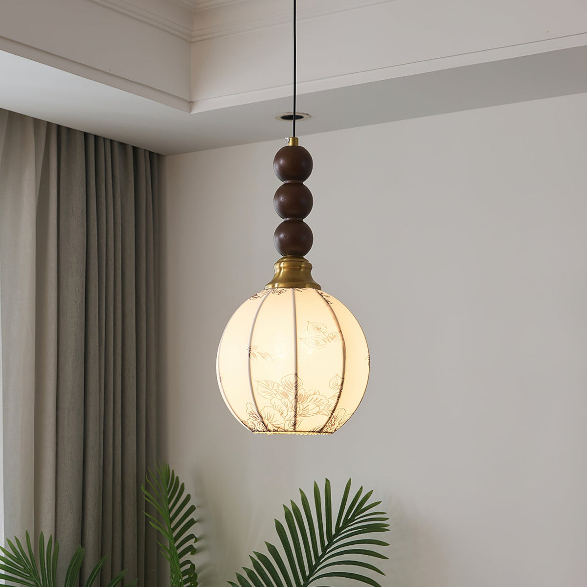 This Clérachic pendant has a round cream shade with fine ribs and a column of three walnut beads, lit warm beside a curtain and a palm.
