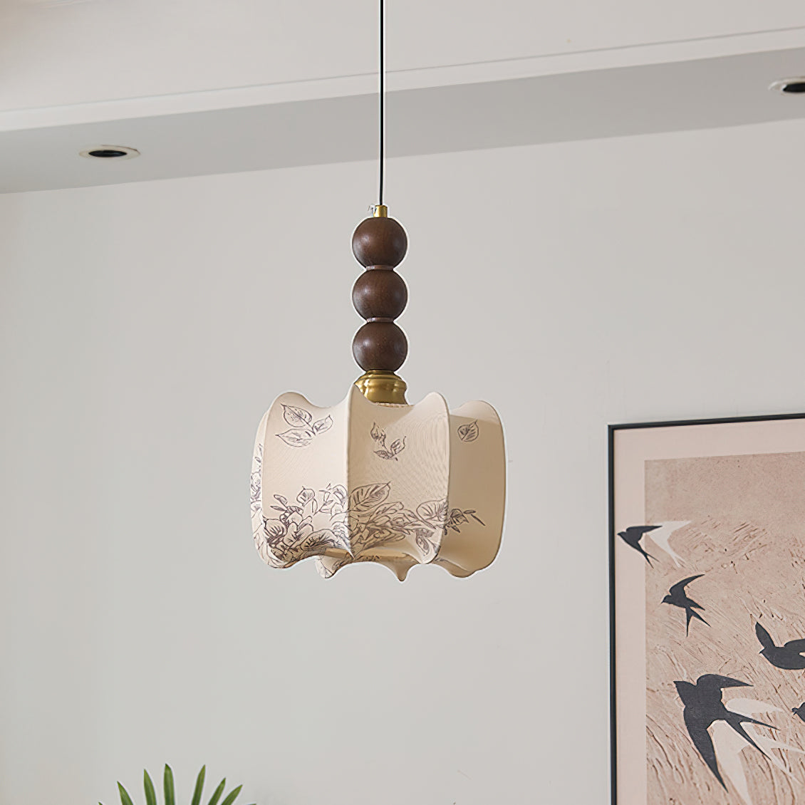 An unlit Clérachic pendant with a pleated cream shade printed with flowers, hung before a framed print of swallows on a pale wall.