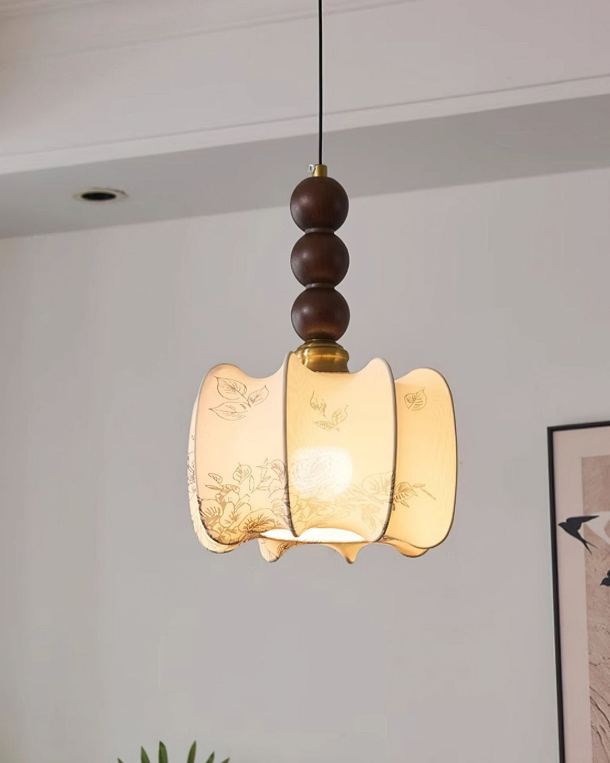 A lit Clérachic pendant with a gathered cream shade and three walnut beads, hung near a ceiling spotlight and a framed bird print.