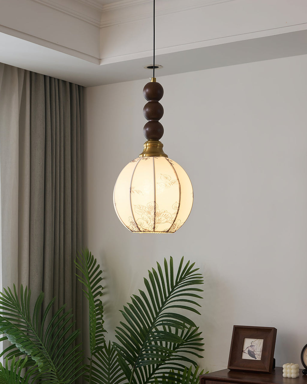 A round-shaded Clérachic pendant with walnut beads, lit warm against a pale wall, with a palm and a framed photograph below.