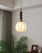 A round-shaded Clérachic pendant with walnut beads, lit warm against a pale wall, with a palm and a framed photograph below.