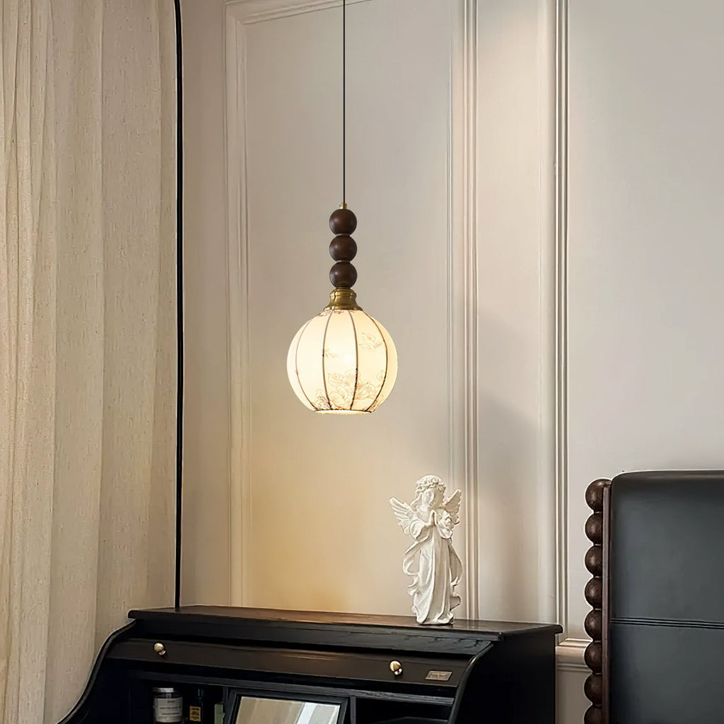 A Clérachic pendant with a round cream shade and walnut beads hangs above a black roll-top desk with an angel figure, beside a curtain.
