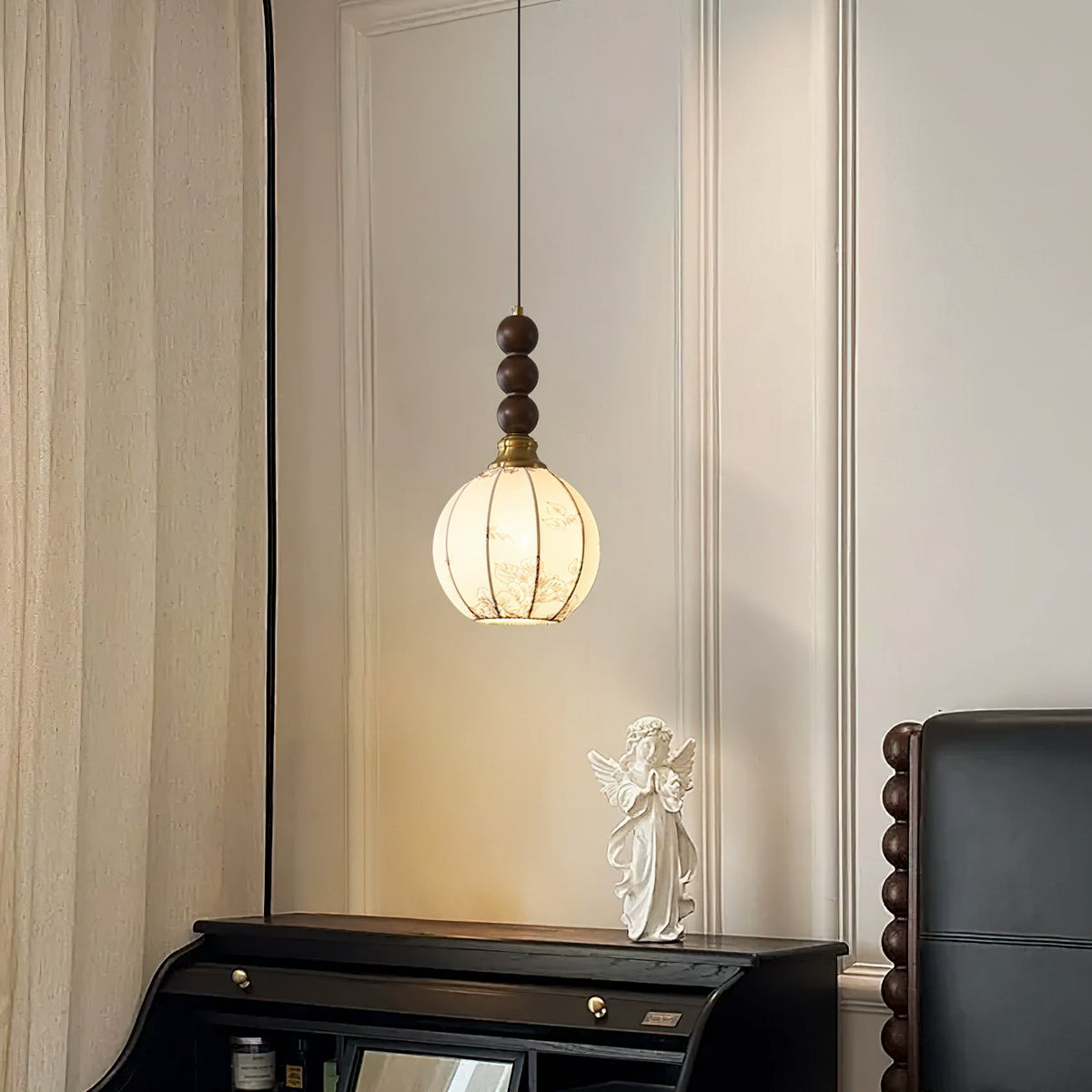 A Clérachic pendant with a round cream shade and walnut beads hangs above a black roll-top desk with an angel figure, beside a curtain.