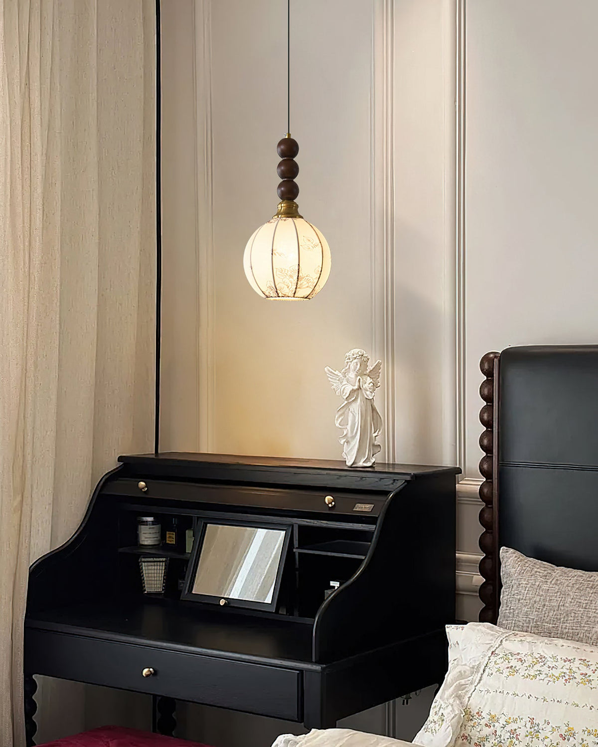 This Clérachic round-shaded pendant hangs over a black desk and an angel figure, beside a carved dark headboard and a cream curtain.