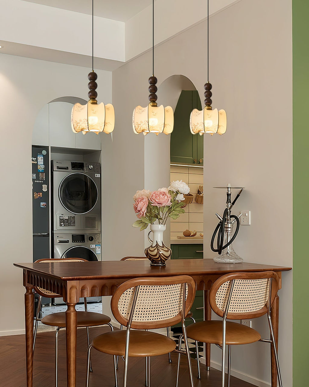 Three Clérachic pendants with gathered cream shades hang above a dining table with cane-backed chairs, near a green arched kitchen.