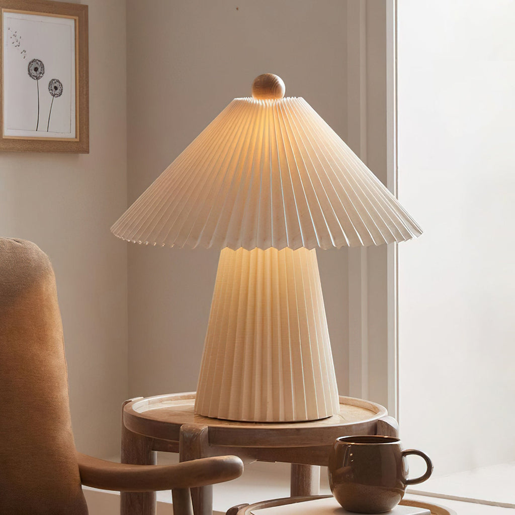 A lit Clérachic table lamp with a pleated beige shade and tapered base on a round wooden side table beside a brown mug and armchair.