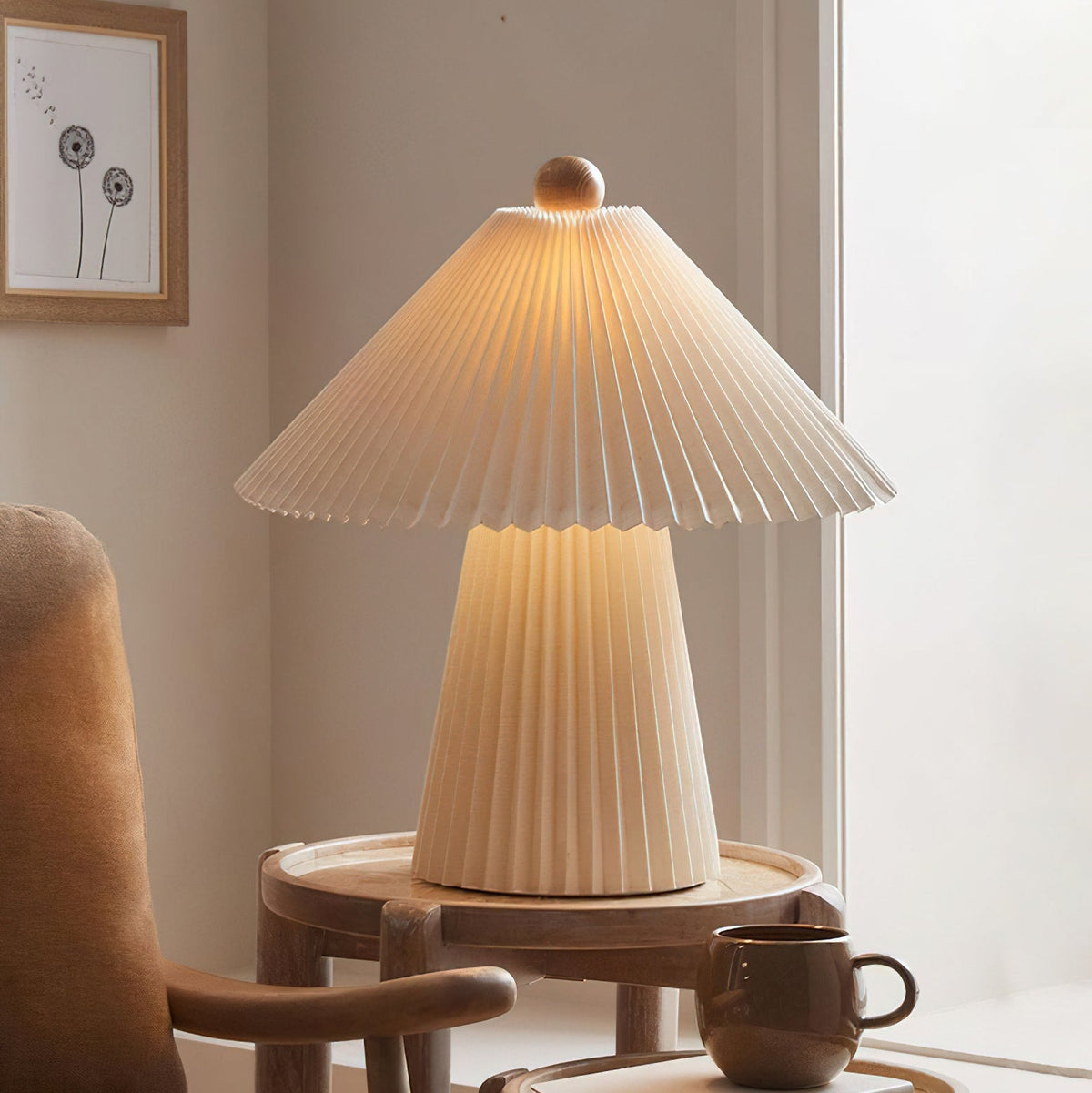 A lit Clérachic table lamp with a pleated beige shade and tapered base on a round wooden side table beside a brown mug and armchair.