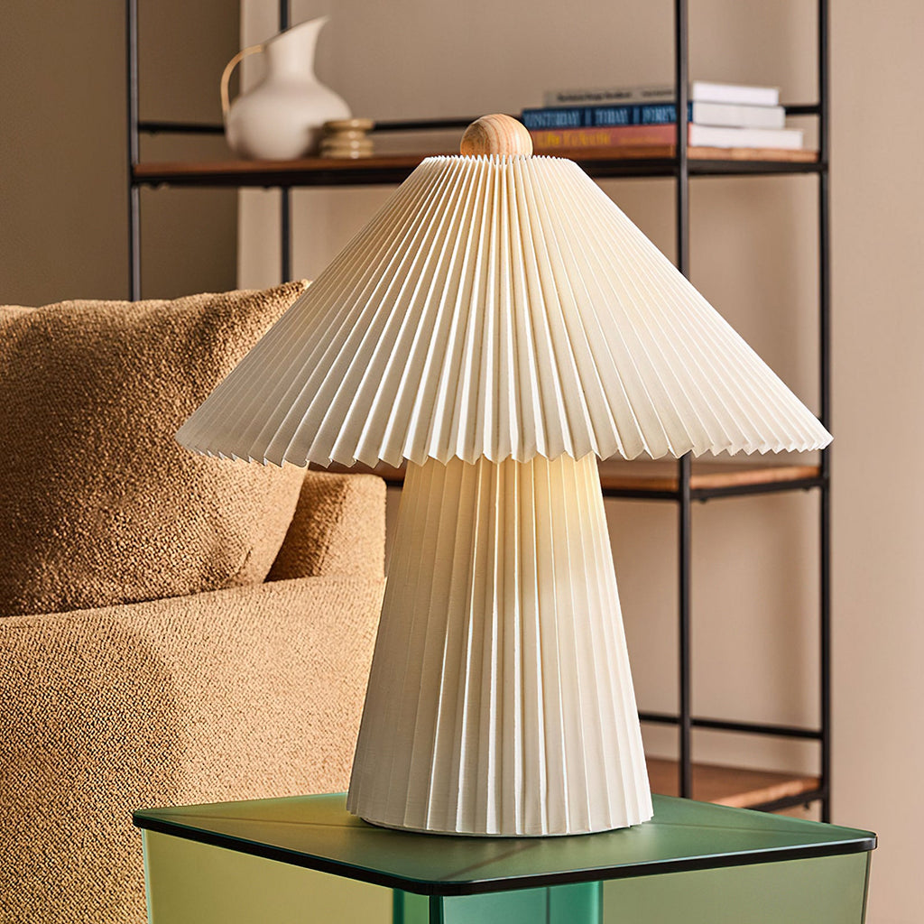 Pleated beige Clérachic table lamp on a green glass side table in front of a shelf unit and a boucle armchair.