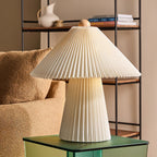 Pleated beige Clérachic table lamp on a green glass side table in front of a shelf unit and a boucle armchair.