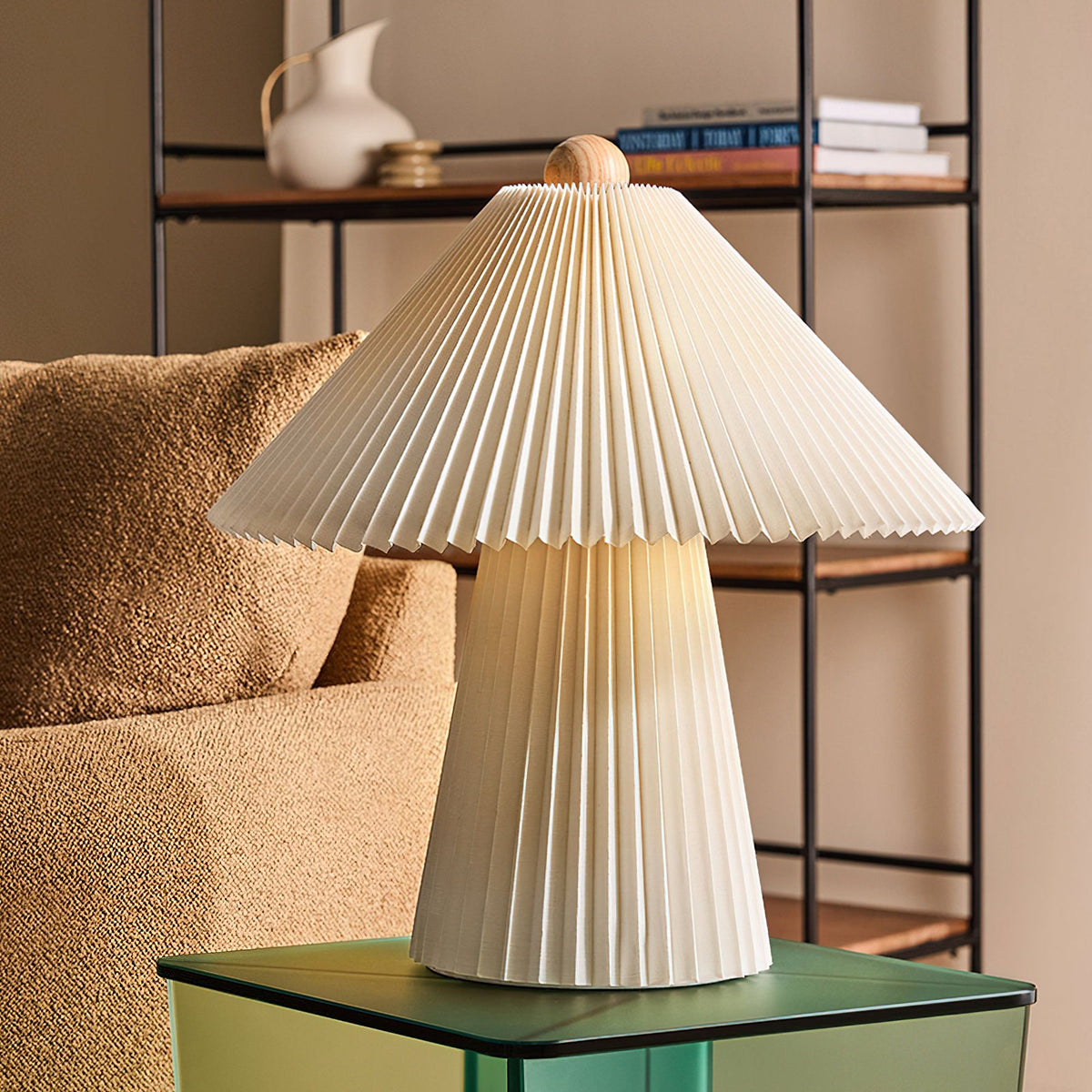 Pleated beige Clérachic table lamp on a green glass side table in front of a shelf unit and a boucle armchair.