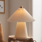 Close view of the lit pleated conical shade and wooden finial of this Clérachic table lamp, above its glowing pleated base.