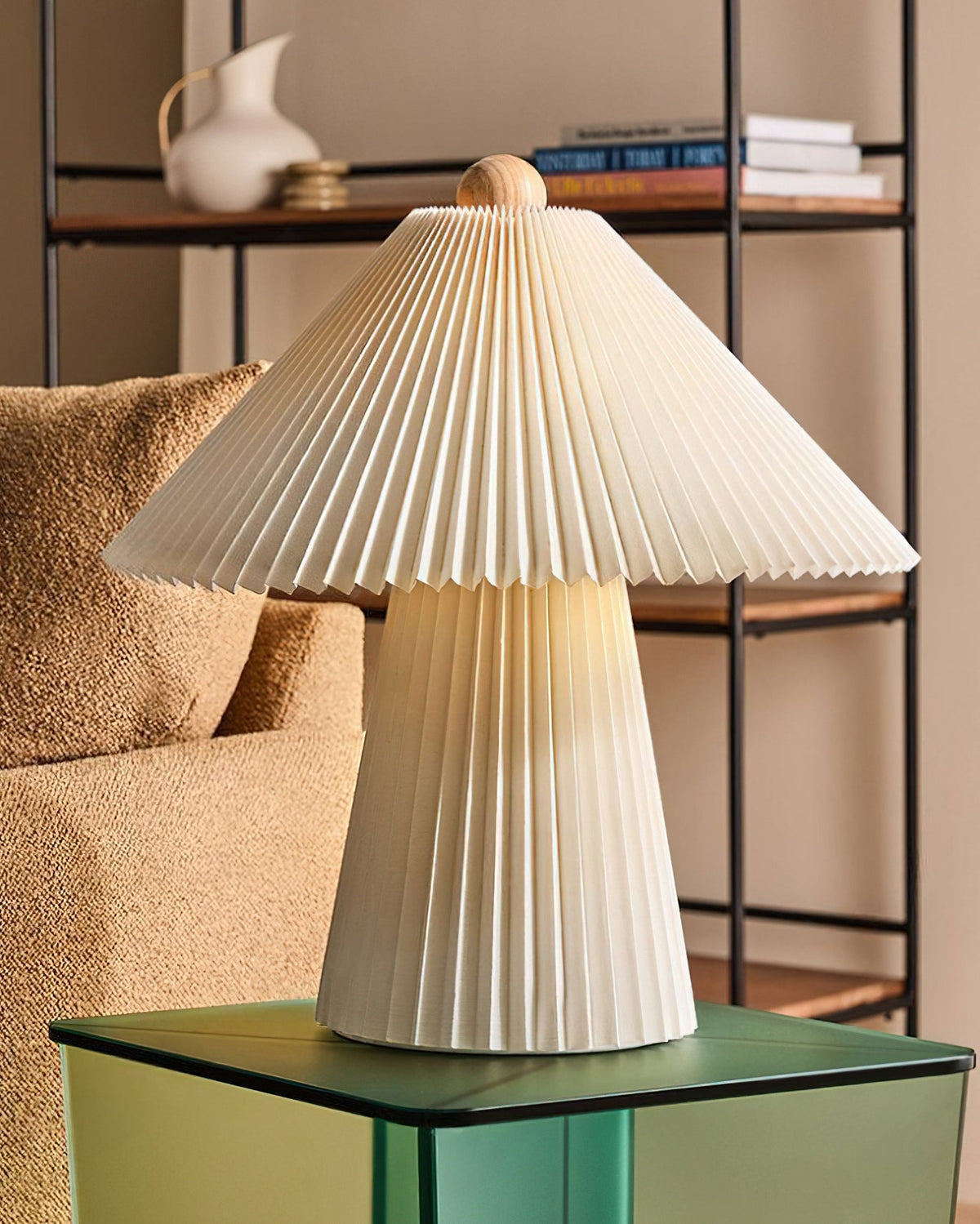 Unlit pleated Clérachic table lamp with a wooden finial on a green glass side table, with a boucle chair and shelf behind.