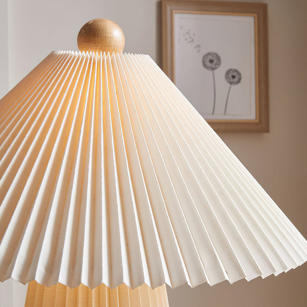 Close view of the pleated shade folds and wooden ball finial of this Clérachic table lamp, lit, with a framed print behind.
