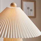 Close view of the pleated shade folds and wooden ball finial of this Clérachic table lamp, lit, with a framed print behind.