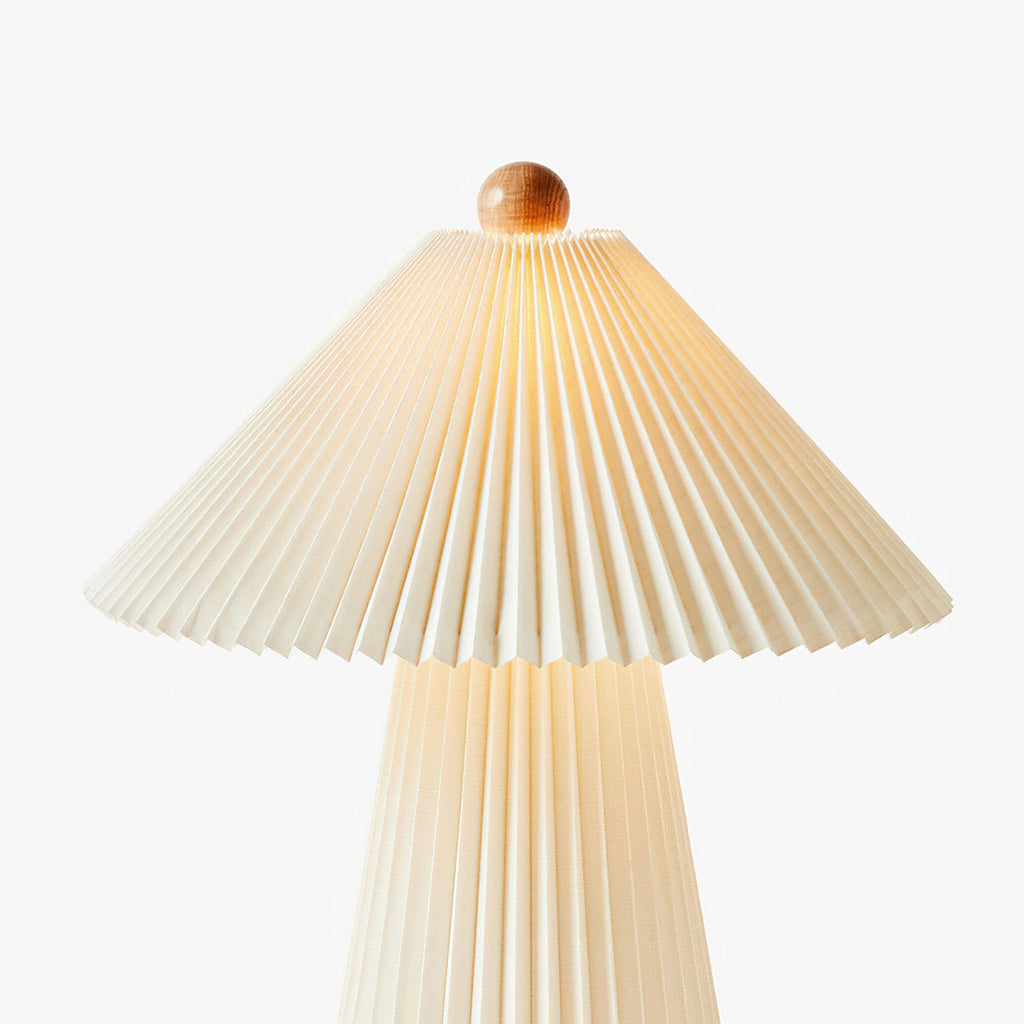 Pleated beige shade with its wooden finial, lit from within, over the top of this Clérachic table lamp on a pale backdrop.