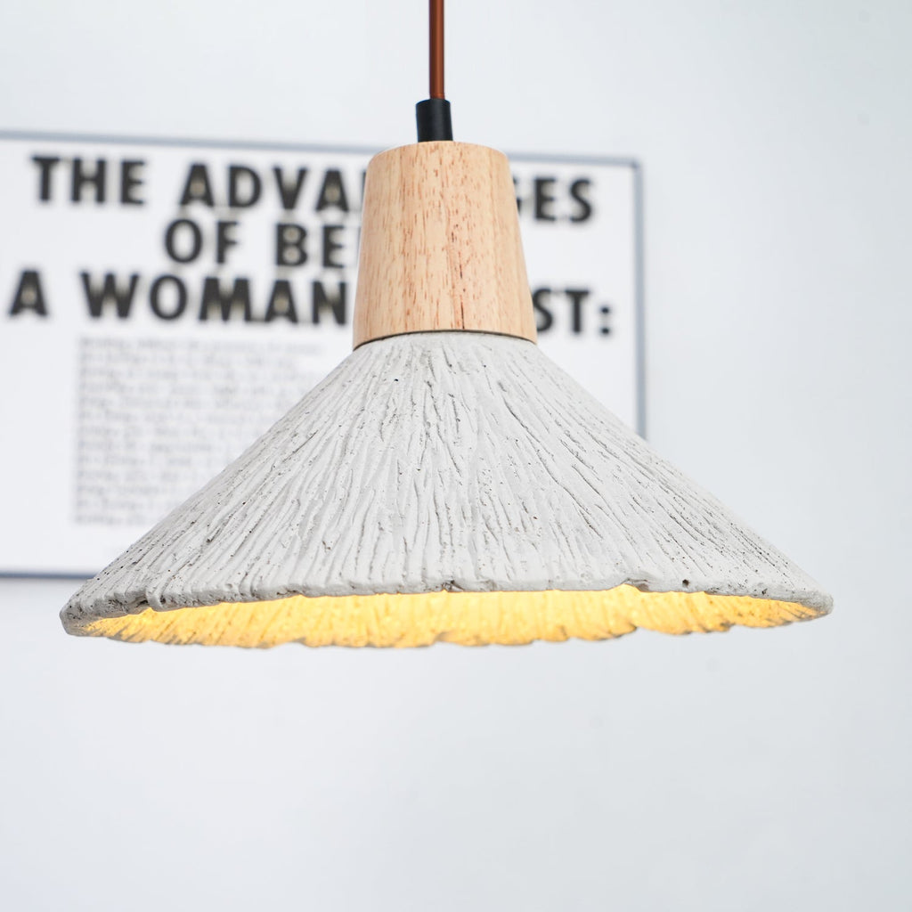 Underside of a wide Clérachic pendant glowing warm, with a blurred framed print behind its wooden cap and ridged grey shade.