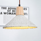 Underside of a wide Clérachic pendant glowing warm, with a blurred framed print behind its wooden cap and ridged grey shade.