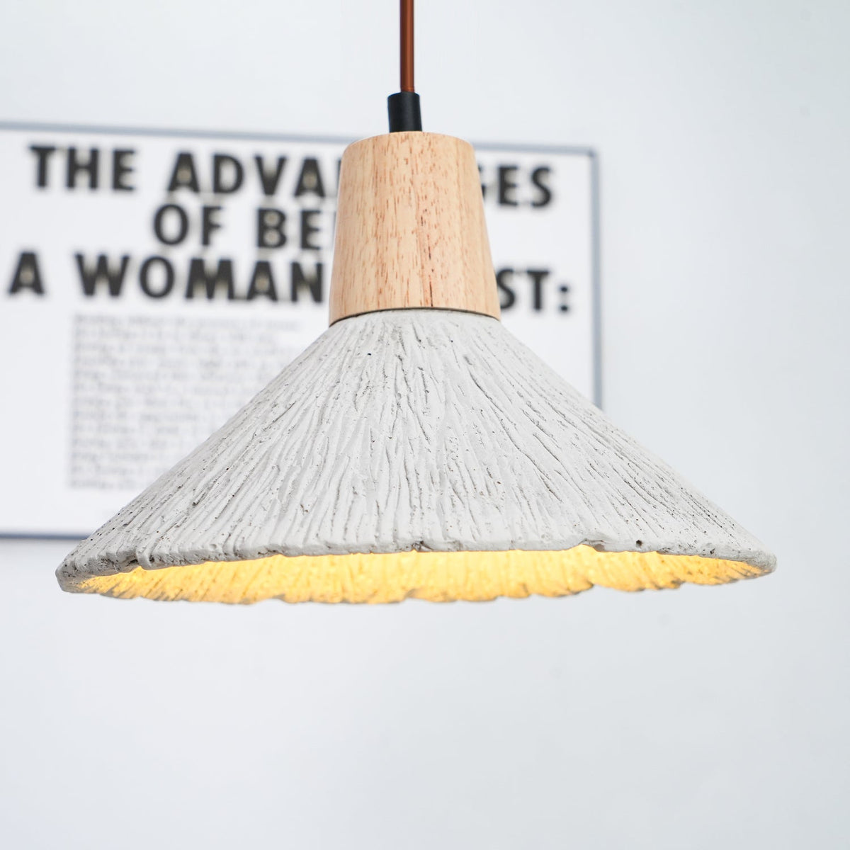 Underside of a wide Clérachic pendant glowing warm, with a blurred framed print behind its wooden cap and ridged grey shade.