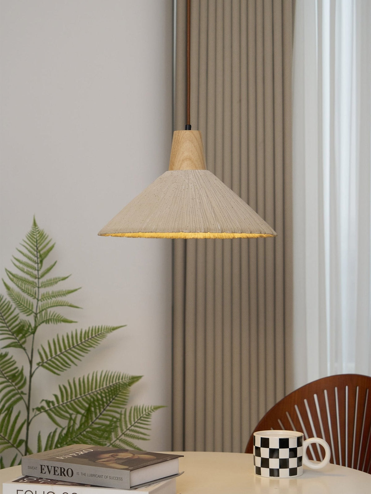 A cream-toned Clérachic pendant hangs beside a fern and striped curtains, over a table with books and a checked mug.