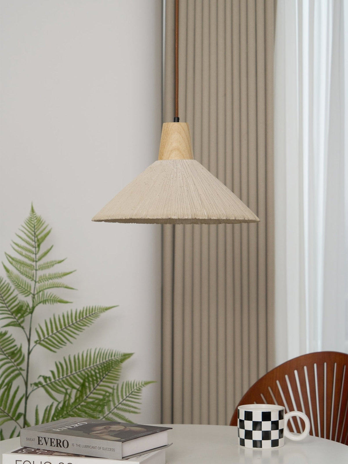 A cream-toned Clérachic pendant, unlit, hangs in front of a striped curtain with a fern and a checked mug on the table below.