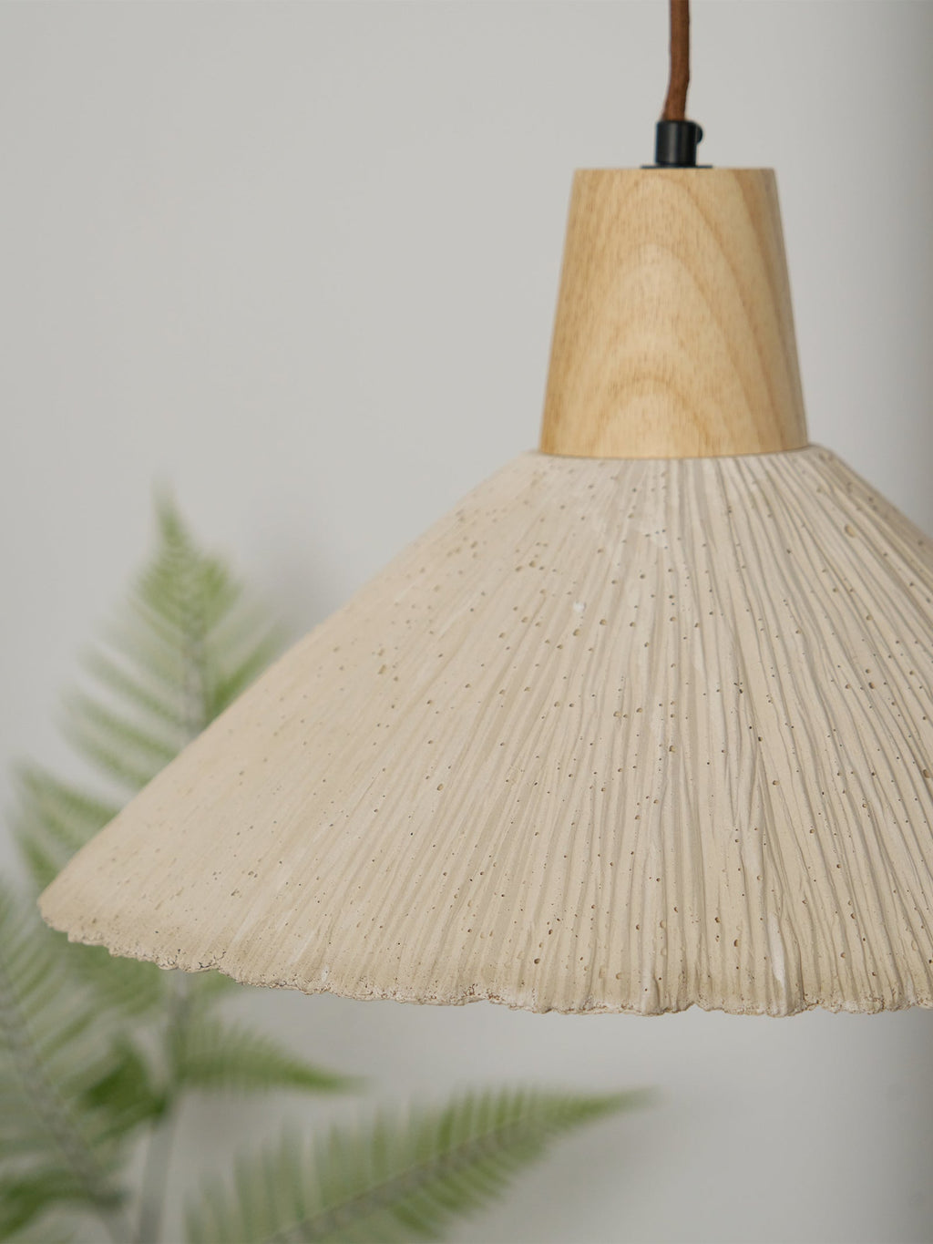 Close view of a cream-toned Clérachic pendant, its fine pleats and wooden cap in focus against a soft green fern.