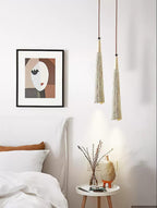 Two slim tapered Clérachic pendants hang by a white bed, next to a framed abstract print and a small round side table.