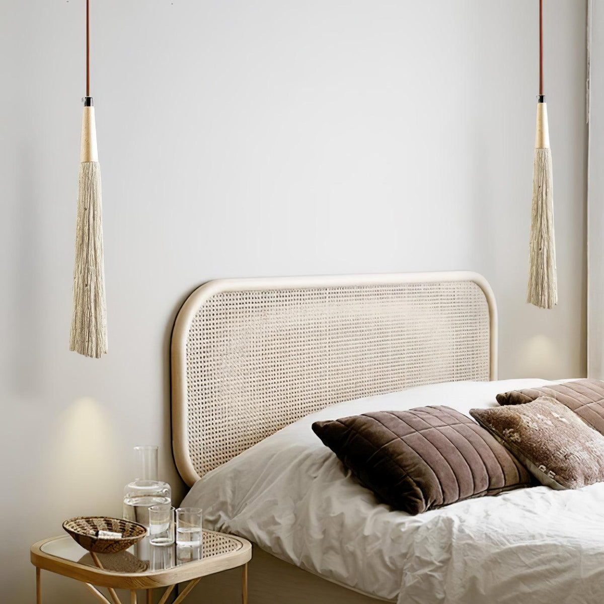 Two long, narrow Clérachic pendants hang above a bed with a rattan headboard and brown cushions.