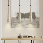 Three narrow Clérachic pendants hang in a row in front of a large landscape painting, above a pale wooden table.