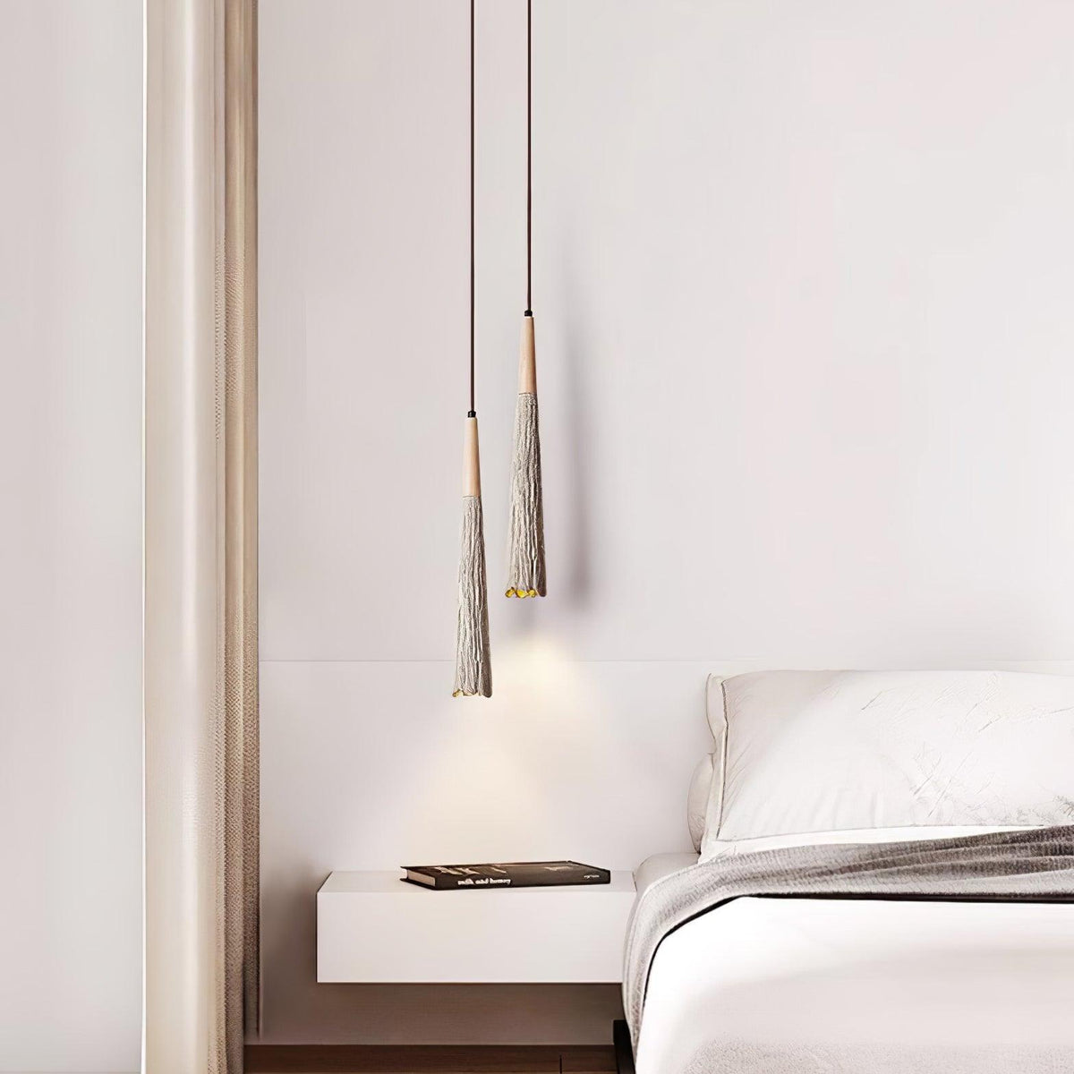 Two slim Clérachic pendants hang at different heights beside a bed, over a floating white bedside shelf with a book.