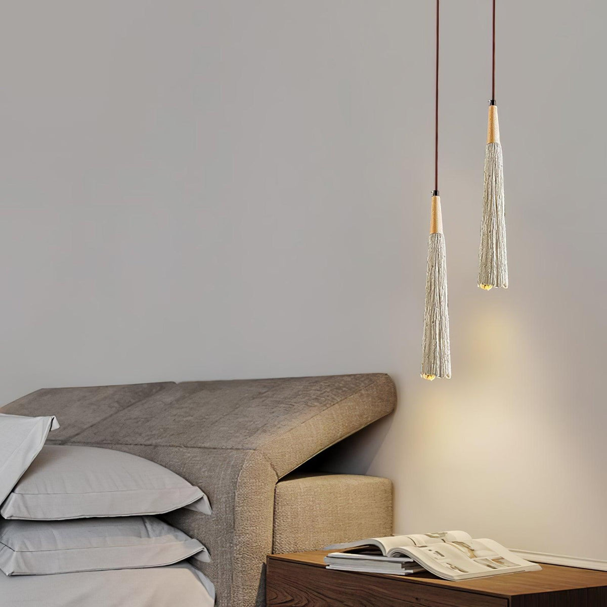 Two slim Clérachic pendants hang above a taupe upholstered headboard, with a magazine on the wooden nightstand below.